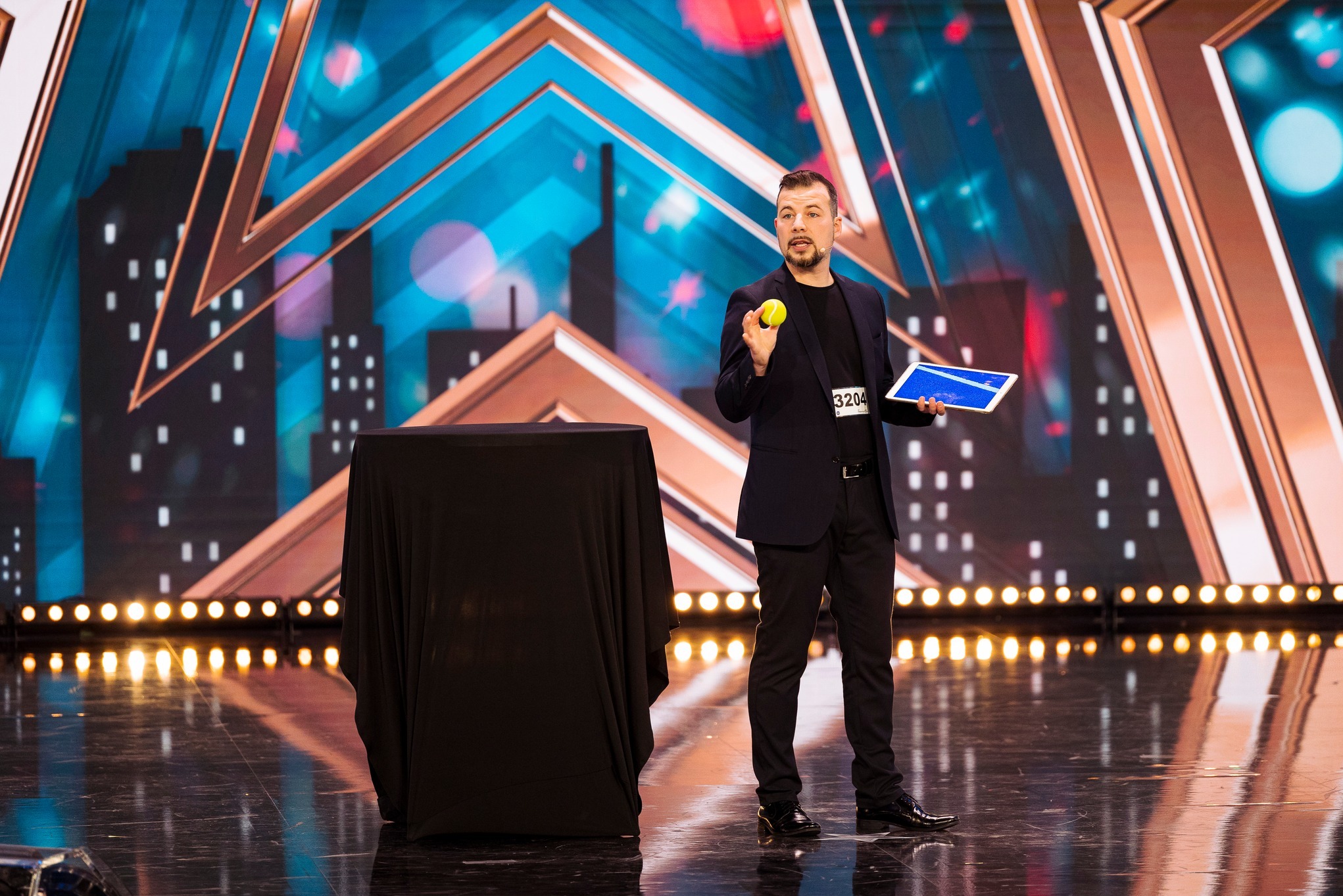 Mężczyzna w czarnym garniturze na scenie z tablet w ręku i żółtą piłeczką, obok stolik przykryty czarnym materiałem, w tle scenografia z motywem miejskim i gwiazdami, casting do programu talent show.