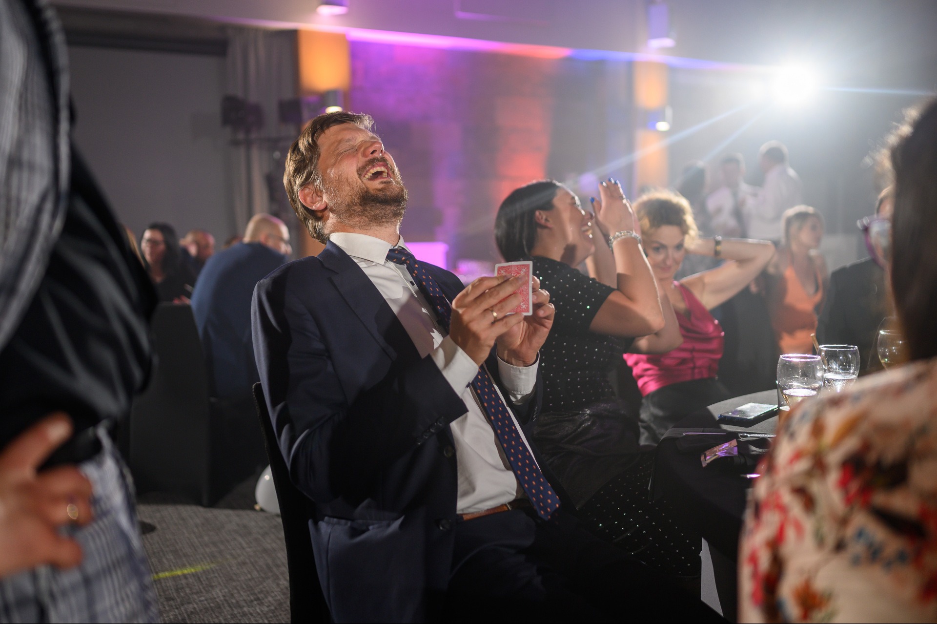 Uśmiechnięty mężczyzna w garniturze trzymający kartę podczas występu na tle publiczności i oświetlenia scenicznego.