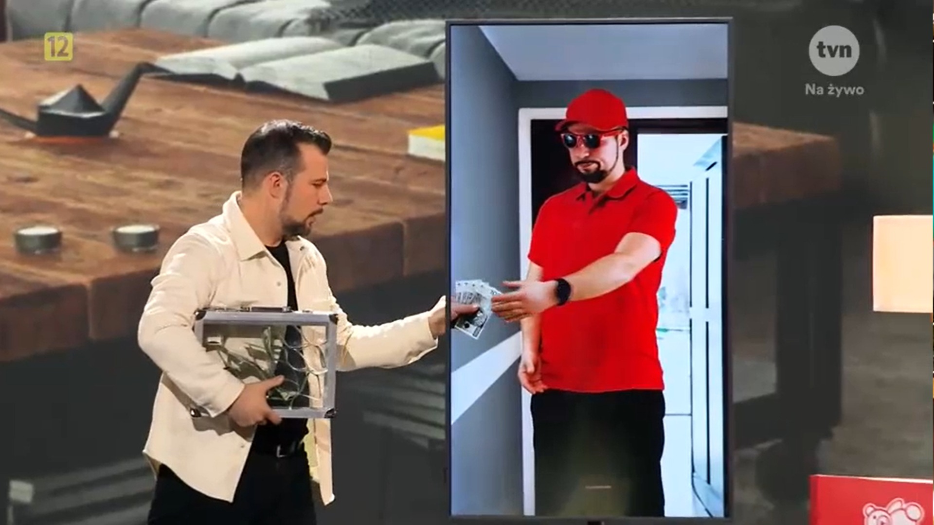 Występ iluzjonisty w telewizji: mężczyzna w jasnej koszuli trzyma metalową walizkę, a na ekranie obok widać mężczyznę w czerwonym stroju, wręczającego banknoty.