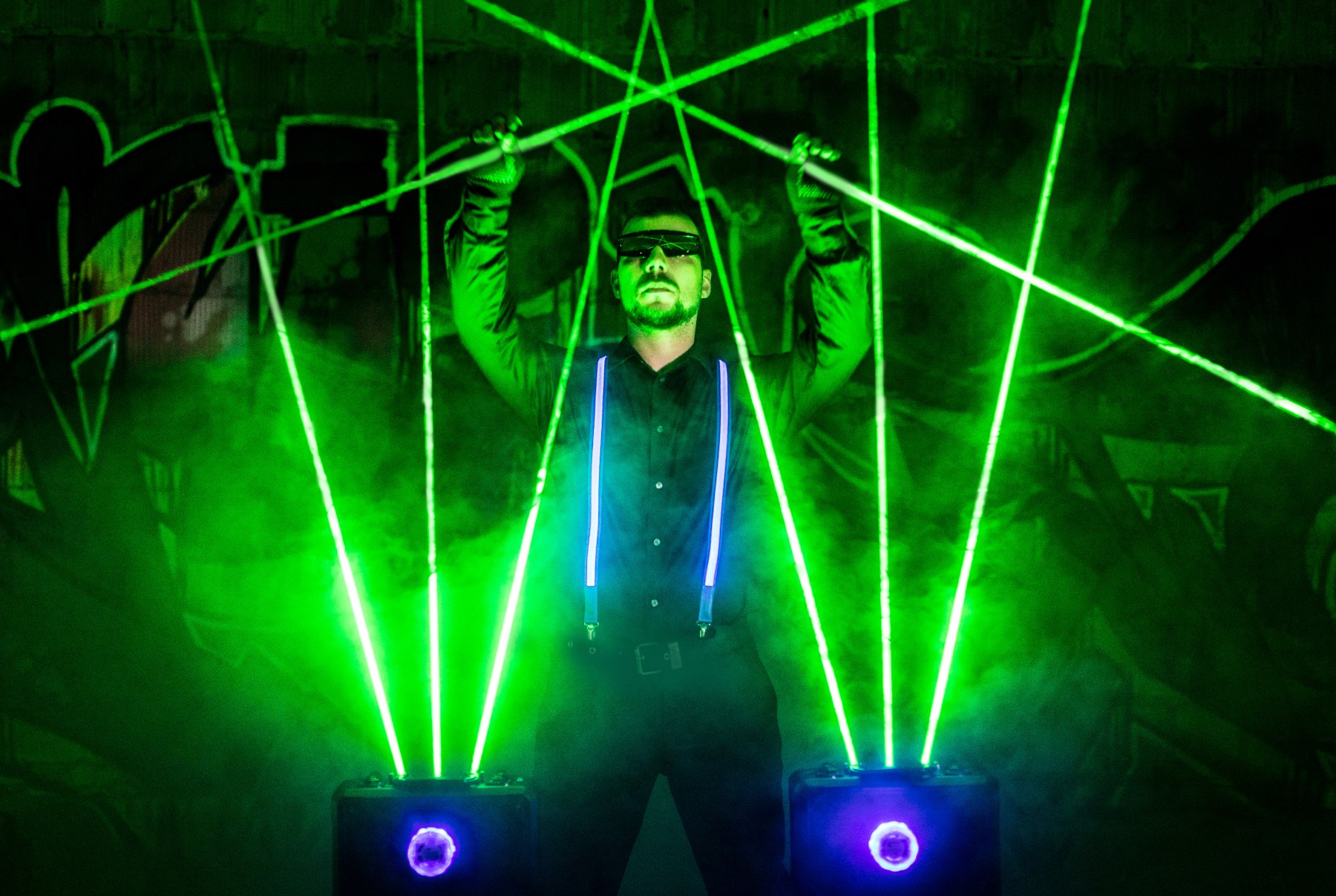 Mężczyzna w okularach przeciwsłonecznych i szelkach, prezentujący pokaz laserowy z zielonymi promieniami na tle graffiti.