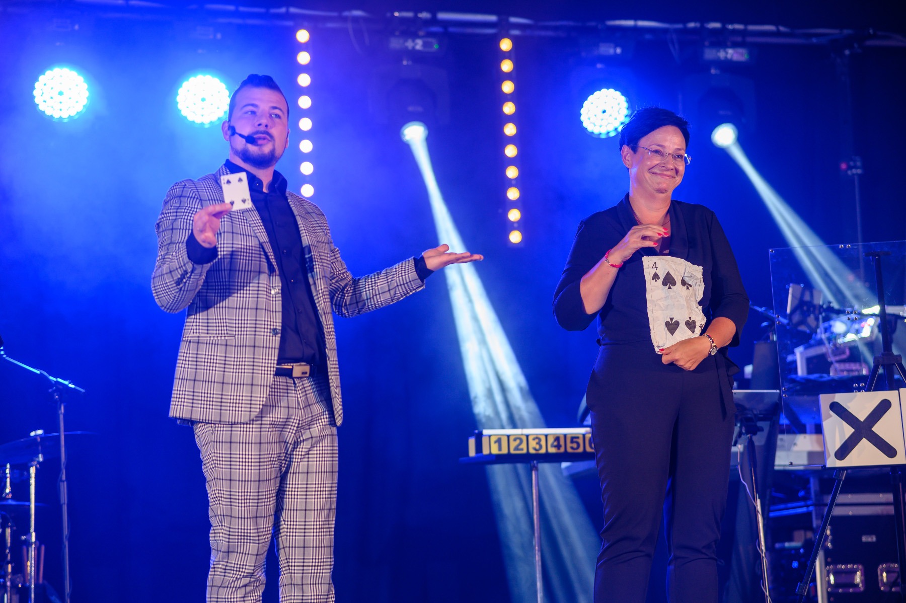 Mężczyzna w kraciastej marynarce i mikrofonem prezentuje kartę do gry, obok kobieta trzyma dużą kartę z czterema pikami na tle oświetlonej sceny.