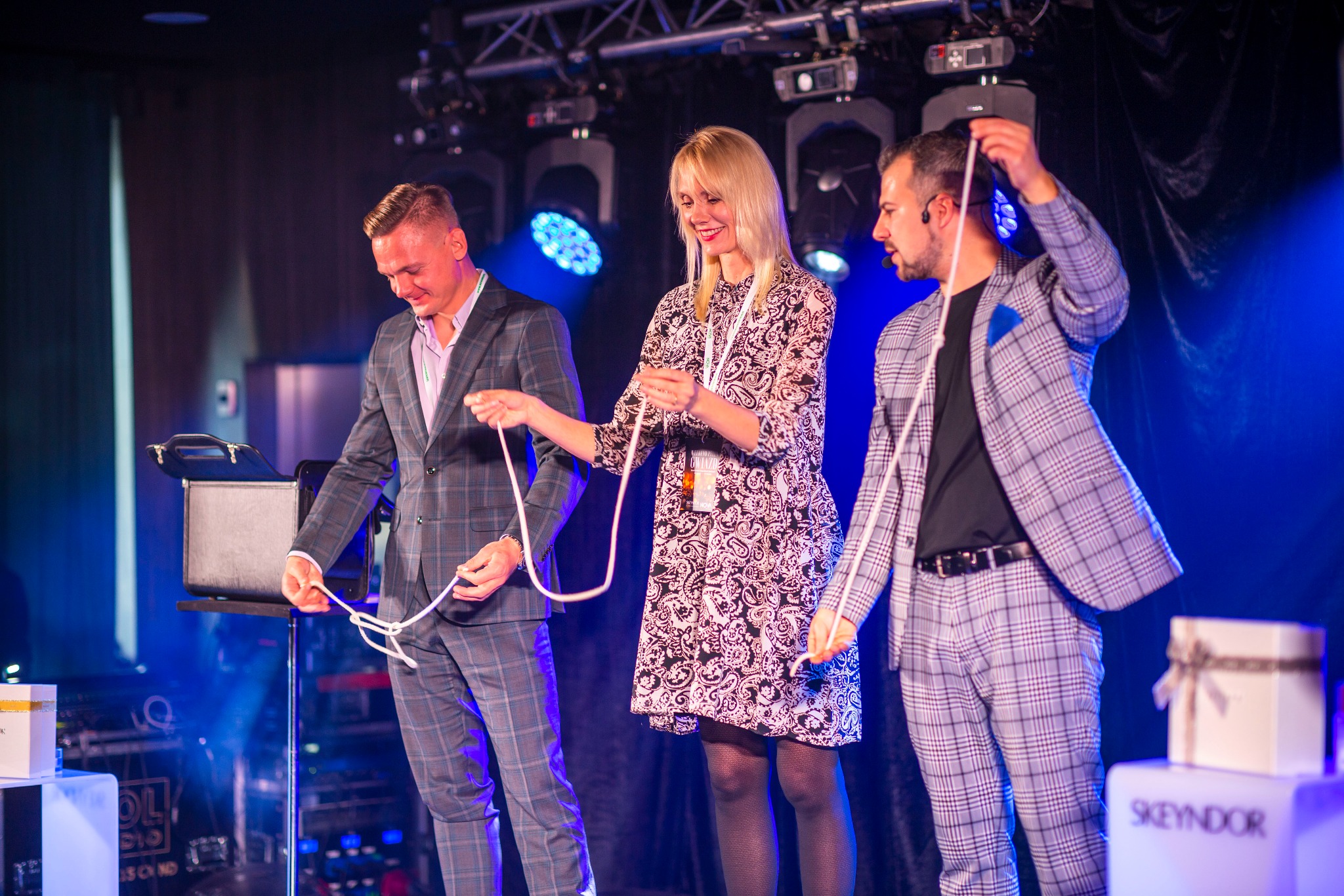 Iluzjonista w kraciastej marynarce i spodniach, kobieta w kwiecistej sukience oraz drugi mężczyzna w podobnym stroju, demonstrują sztuczkę z liną na scenie oświetlonej niebieskim światłem, w tle...