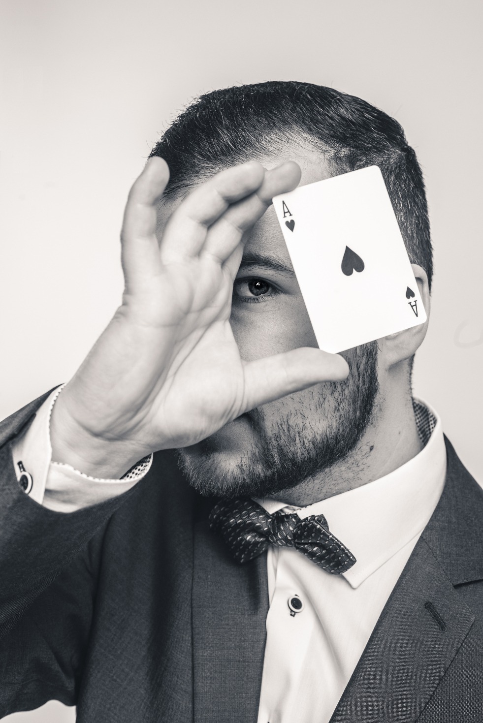 Czarno-białe zbliżenie mężczyzny w garniturze i muszce, zasłaniającego oko asem pik, widoczne mankiety koszuli z ozdobnymi spinkami.