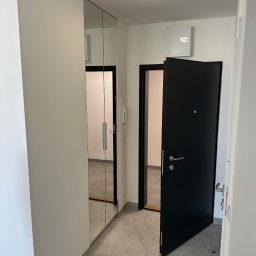 MONTECH SPÓŁKA Z OGRANICZONĄ ODPOWIEDZIALNOŚCIĄ - Minimalistyczny przedpokój: szara podłoga, białe ściany, lustrzana szafa wnękowa i czarne drzwi wejściowe, z widocznym fragmentem klimatyzacji na ścianie.