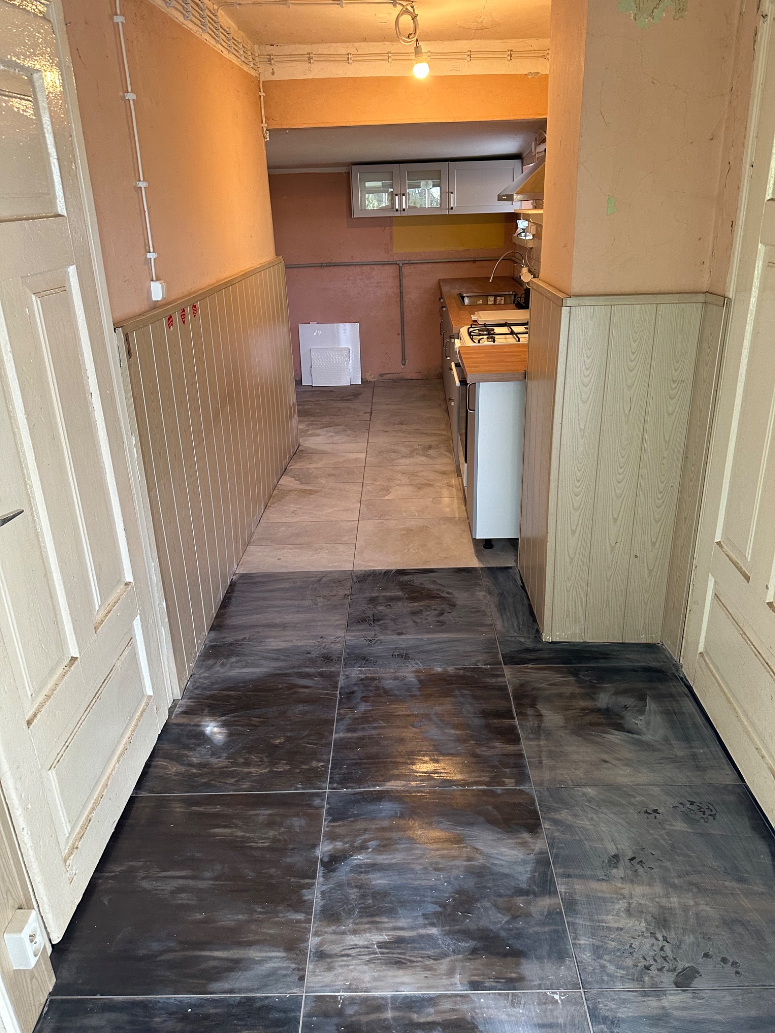 Korytarz z ciemnymi, błyszczącymi płytkami podłogowymi, przechodzący w jaśniejsze płytki w kierunku kuchni. Ściany wykończone boazerią i pomalowane na beżowo.