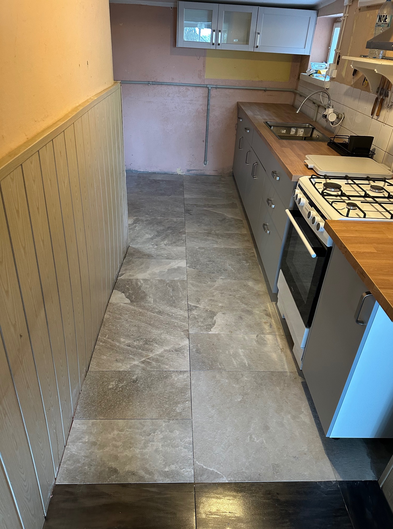 Szare płytki podłogowe z wzorem marmuru w kuchni z szarą zabudową i drewnianym blatem. Widoczny fragment ściany z boazerią i fragmenty instalacji elektrycznej.