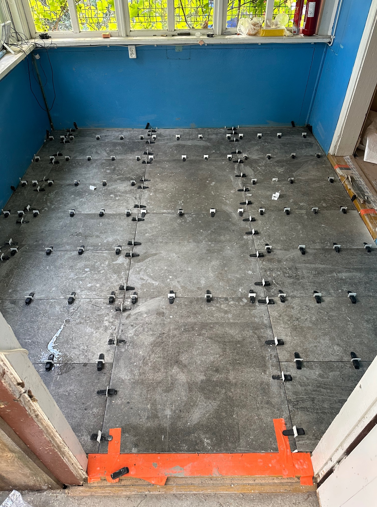 Szare płytki podłogowe w trakcie układania, z użyciem klipsów poziomujących. Niebieska ściana i okno w tle. Widoczny fragment progu z pomarańczową listwą.