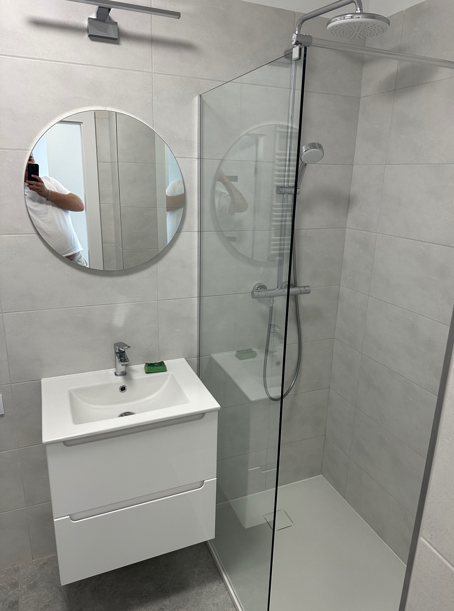 Nowoczesna łazienka w szarościach: kabina prysznicowa typu walk-in, okrągłe lustro z odbiciem osoby robiącej zdjęcie, biała szafka z umywalką, minimalistyczne oświetlenie.