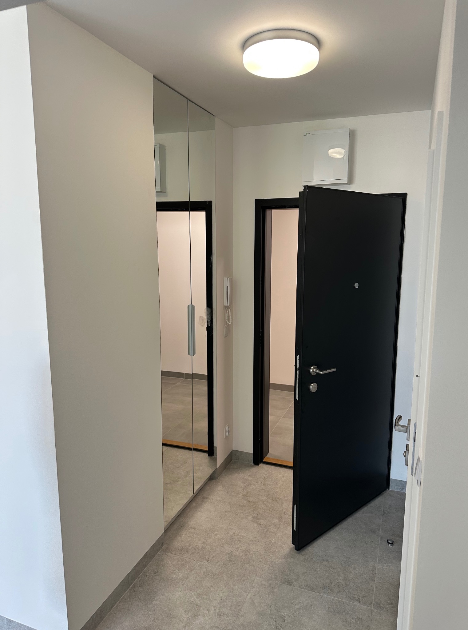 Minimalistyczny przedpokój: szara podłoga, białe ściany, lustrzana szafa wnękowa i czarne drzwi wejściowe, z widocznym fragmentem klimatyzacji na ścianie.