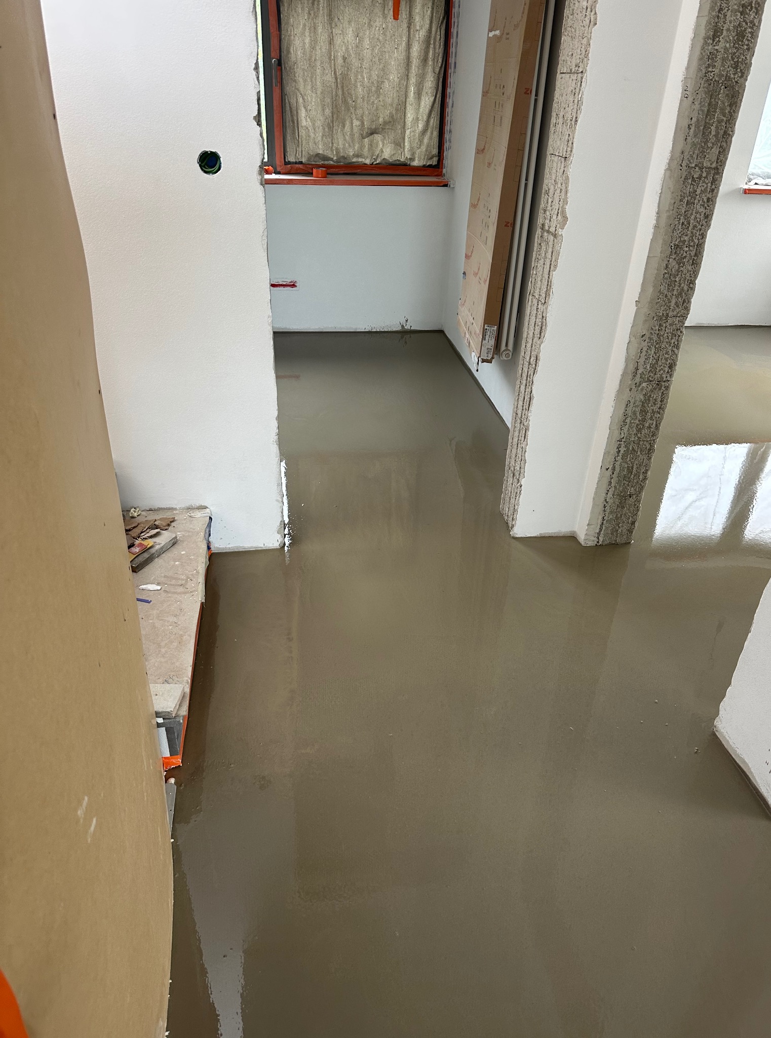 Wylany, mokry podkład podłogowy w jasnym pomieszczeniu z oknami. Widoczne narzędzia na krawędzi podłoża. Świeżo wylana wylewka podłogowa.