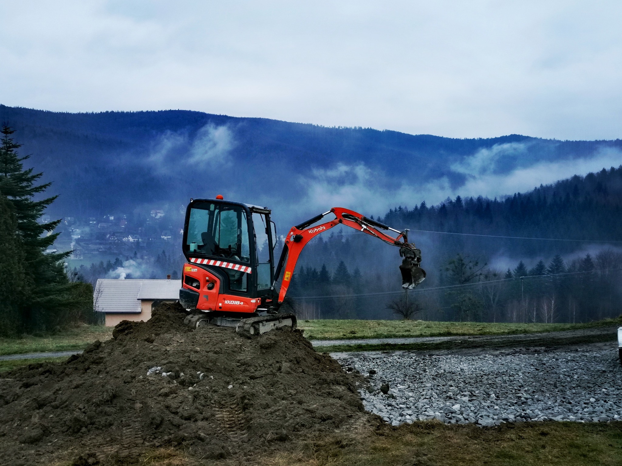 Pomarańczowa mini-koparka Kubota KX018-4 na pryzmie ziemi, w tle widok na góry spowite mgłą i fragment utwardzonej drogi żwirowej.