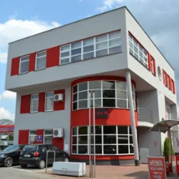 Nowoczesny, trzypiętrowy budynek stacji benzynowej i myjni samochodowej z dużymi, przeszklonymi oknami, pomalowany w szaro-czerwone barwy, widoczny z zewnątrz, z zaparkowanymi samochodami.