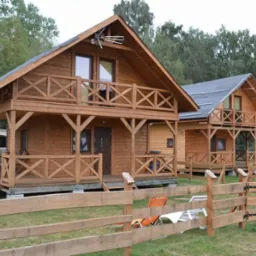 Dwa drewniane domki letniskowe z balkonami i drewnianą balustradą, ogrodzone niskim płotem na tle zieleni.