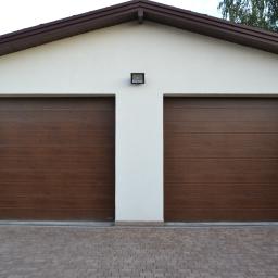 Tom-Bud Tomasz Nowak - Dwie brązowe segmentowe bramy garażowe w jasnym budynku z brązowym dachem i kostką brukową na podjeździe.