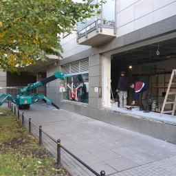 Tom-Bud Tomasz Nowak - Montaż okna wystawowego w lokalu usługowym przy użyciu specjalistycznego dźwigu pająka; dwóch monterów pracuje wewnątrz, obok stoi drabina, a za oknem widać drzewo z żółknącymi liśćmi.