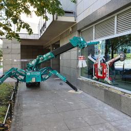 Tom-Bud Tomasz Nowak - Montaż okna w budynku biurowym w Żurominie przy użyciu specjalistycznego dźwigu z przyssawkami; widoczne zabezpieczenia i częściowo zamontowane okno.