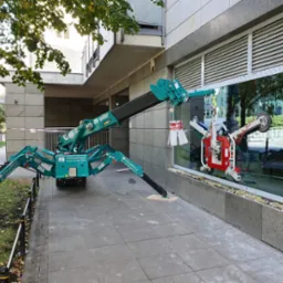 Montaż okna w budynku biurowym w Żurominie przy użyciu specjalistycznego dźwigu z przyssawkami; widoczne zabezpieczenia i częściowo zamontowane okno.