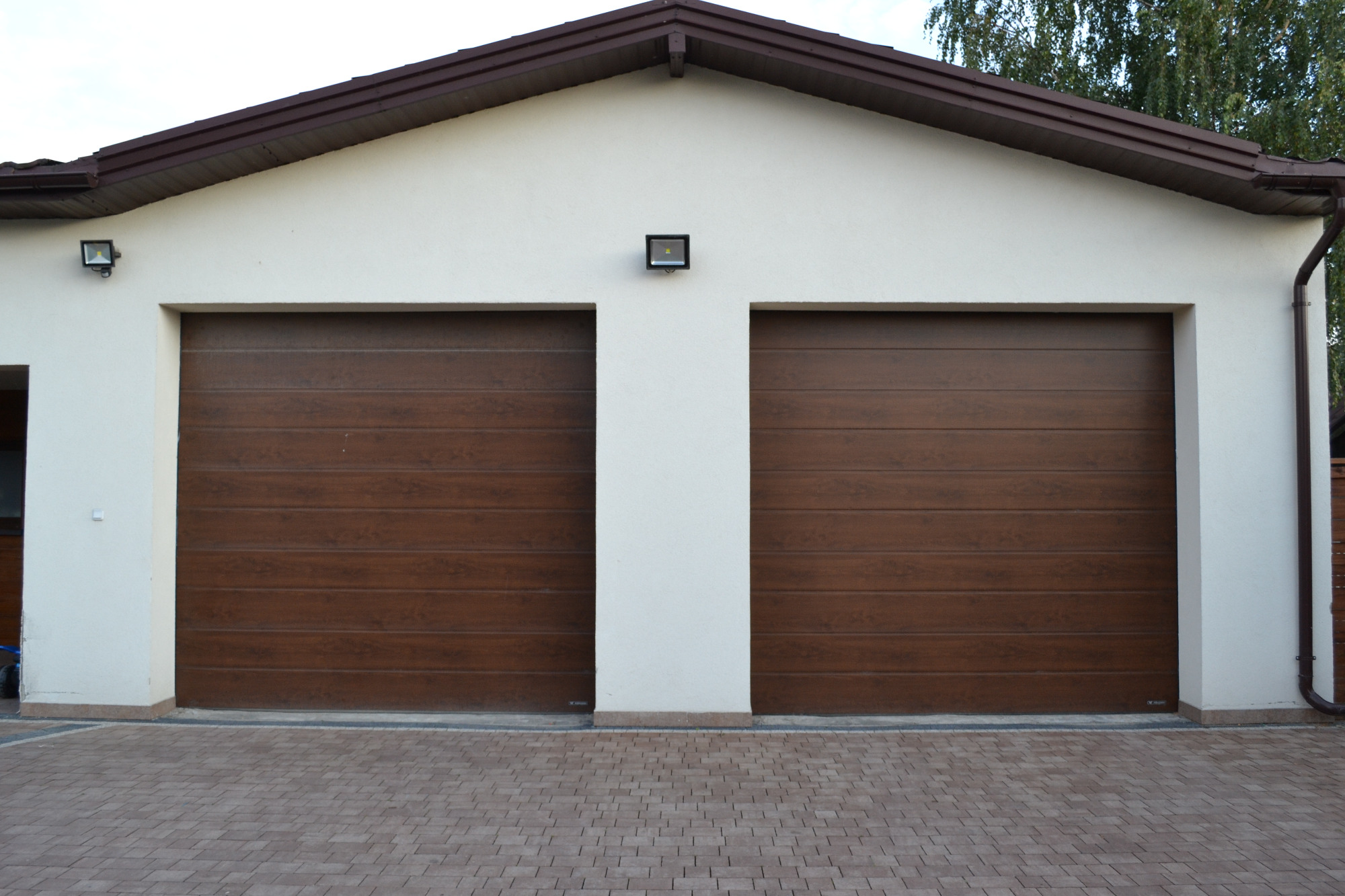 Dwie brązowe segmentowe bramy garażowe w jasnym budynku z brązowym dachem i kostką brukową na podjeździe.