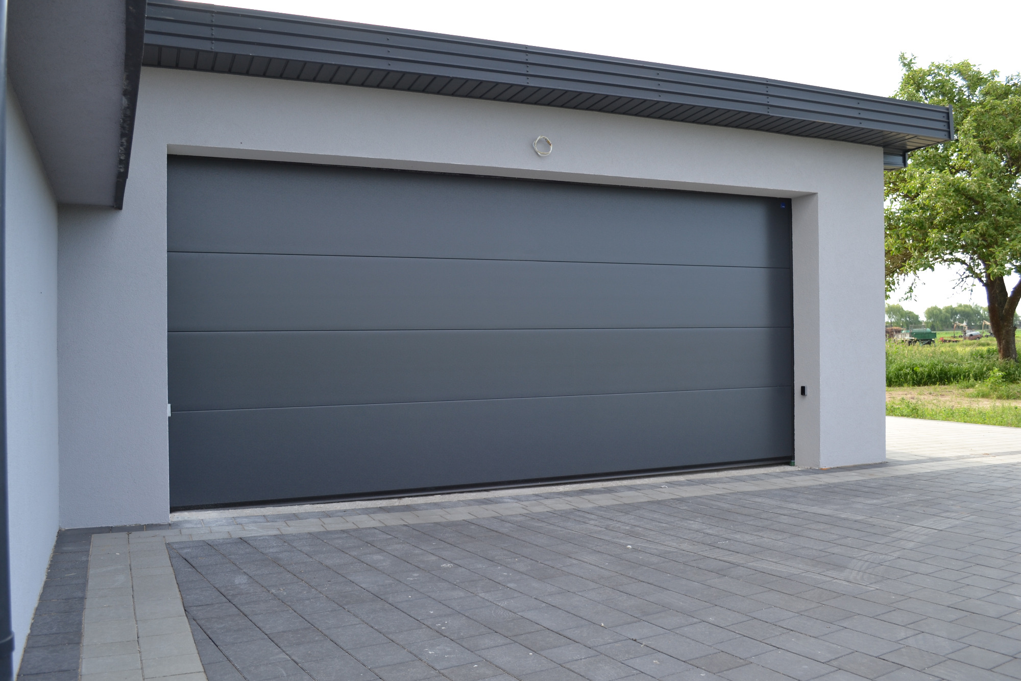 Szara, segmentowa brama garażowa z poziomymi panelami, zamontowana w nowoczesnym garażu z szarą elewacją i kostką brukową na podjeździe, widok z zewnątrz.