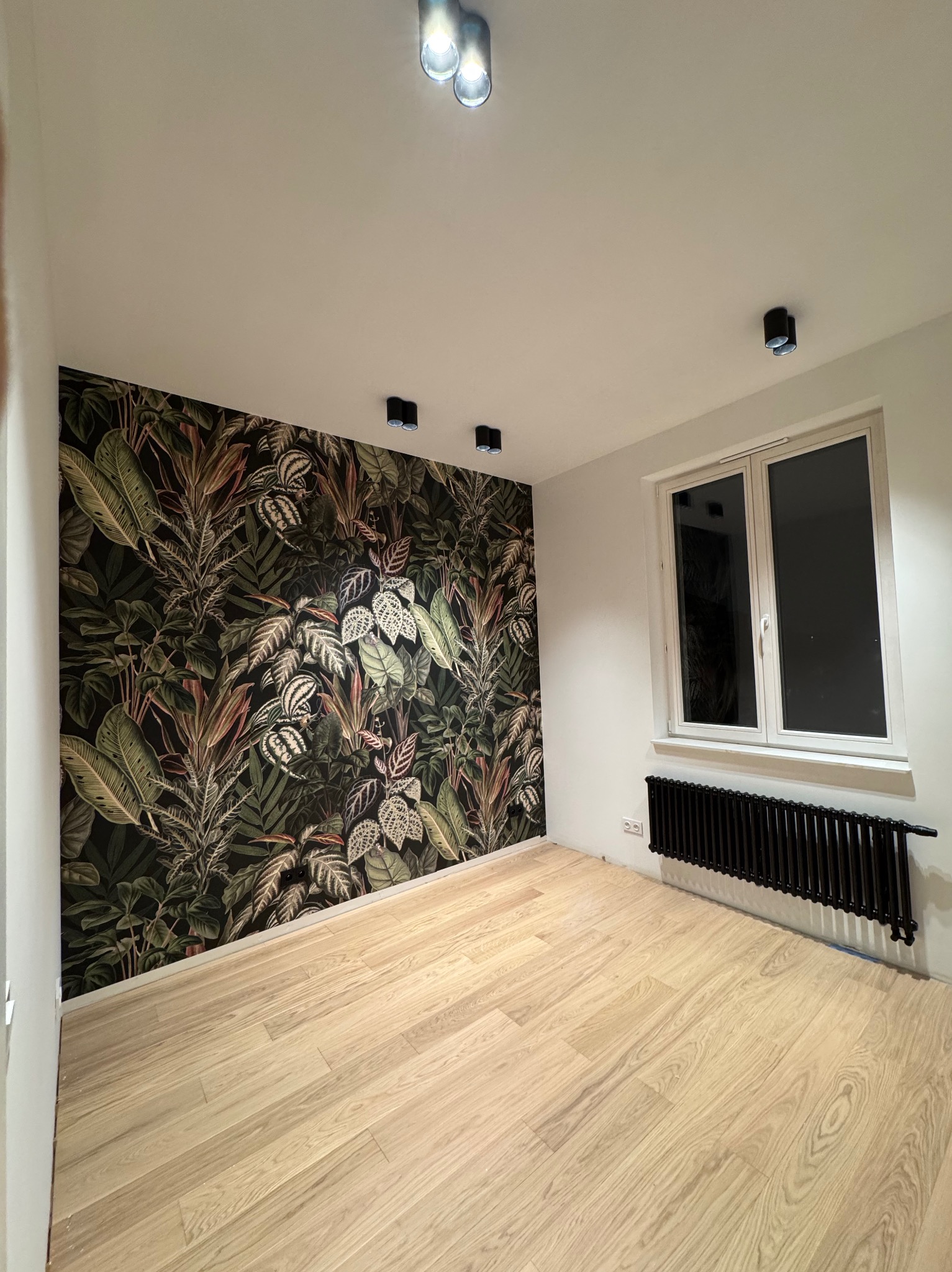 Pokój po remoncie z drewnianą podłogą, ścianą z tapetą w motywy roślinne i czarnym grzejnikiem pod oknem, oświetlony przez reflektory sufitowe.