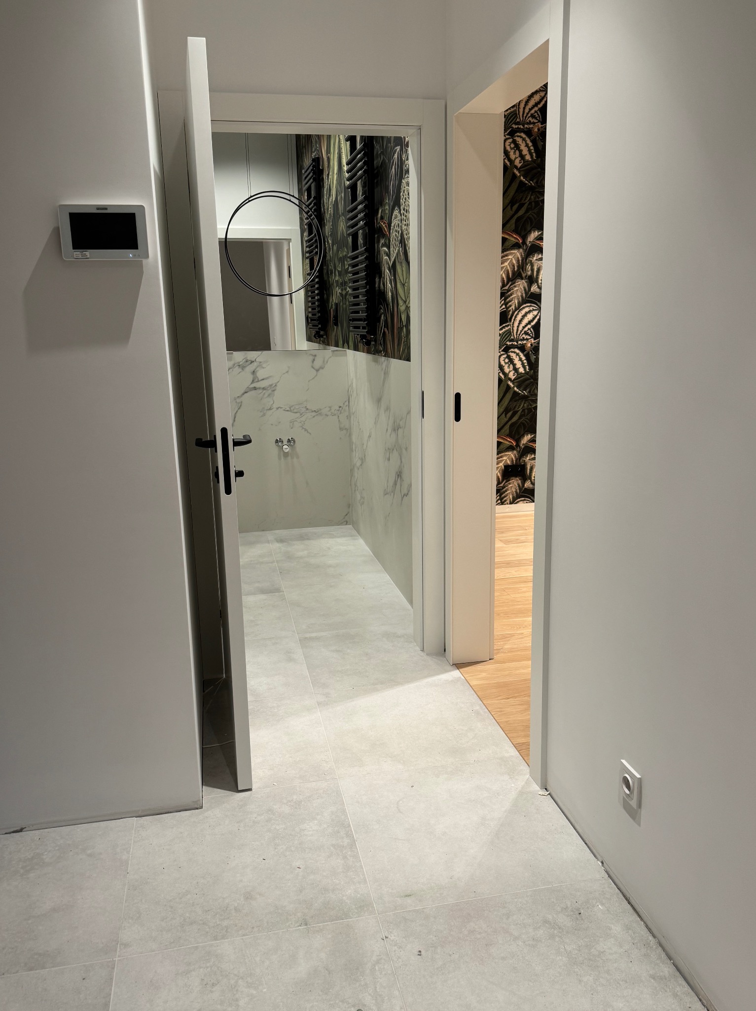 Nowoczesne wnętrze z otwartymi drzwiami do łazienki z marmurową ścianą i okrągłym lustrem oraz pokoju z tapetą w motywy roślinne, widok z korytarza z szarą podłogą.