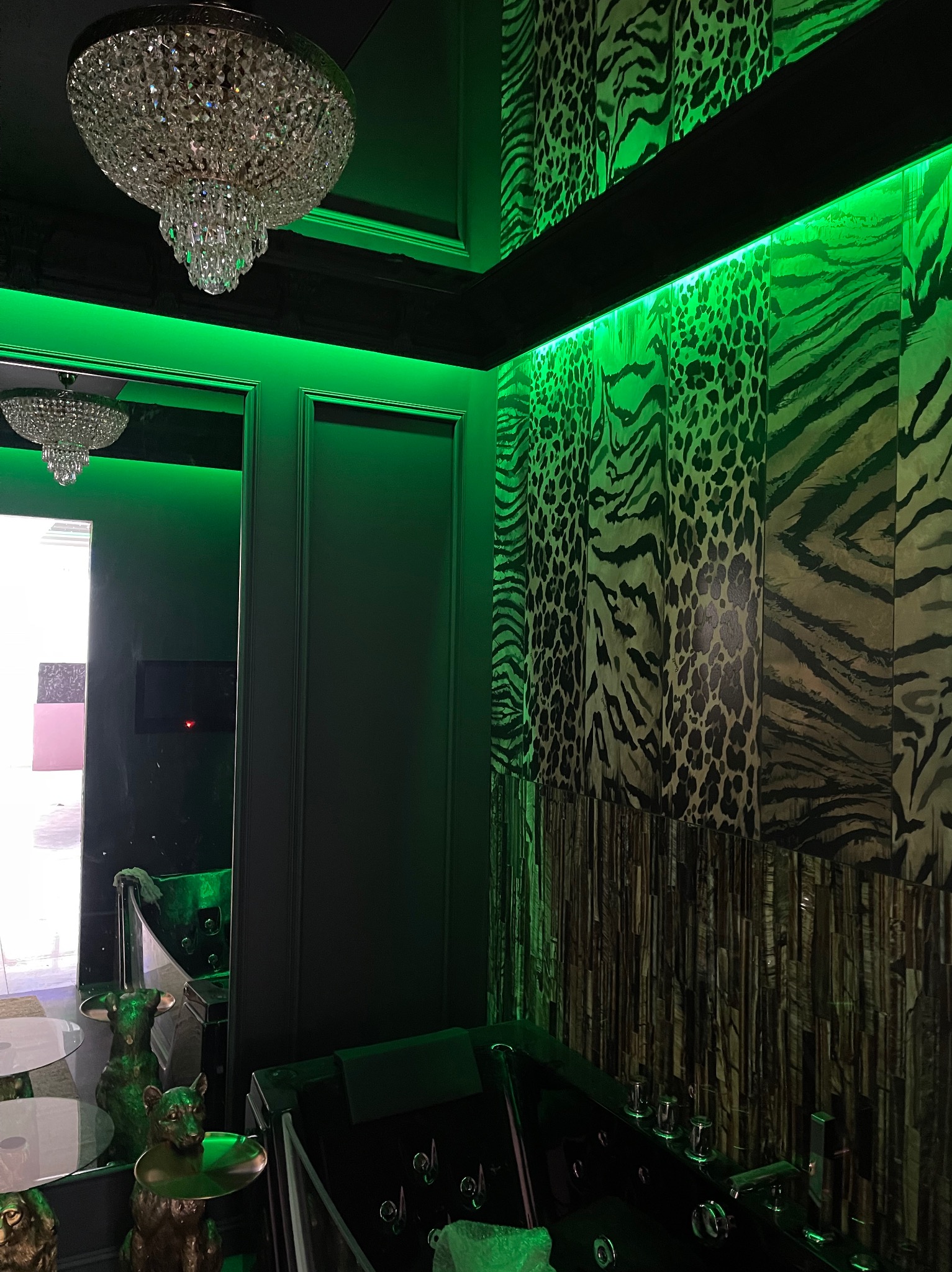 Luksusowa łazienka z czarną wanną z hydromasażem, ściany w zwierzęce wzory, kryształowy żyrandol i zielone oświetlenie LED.