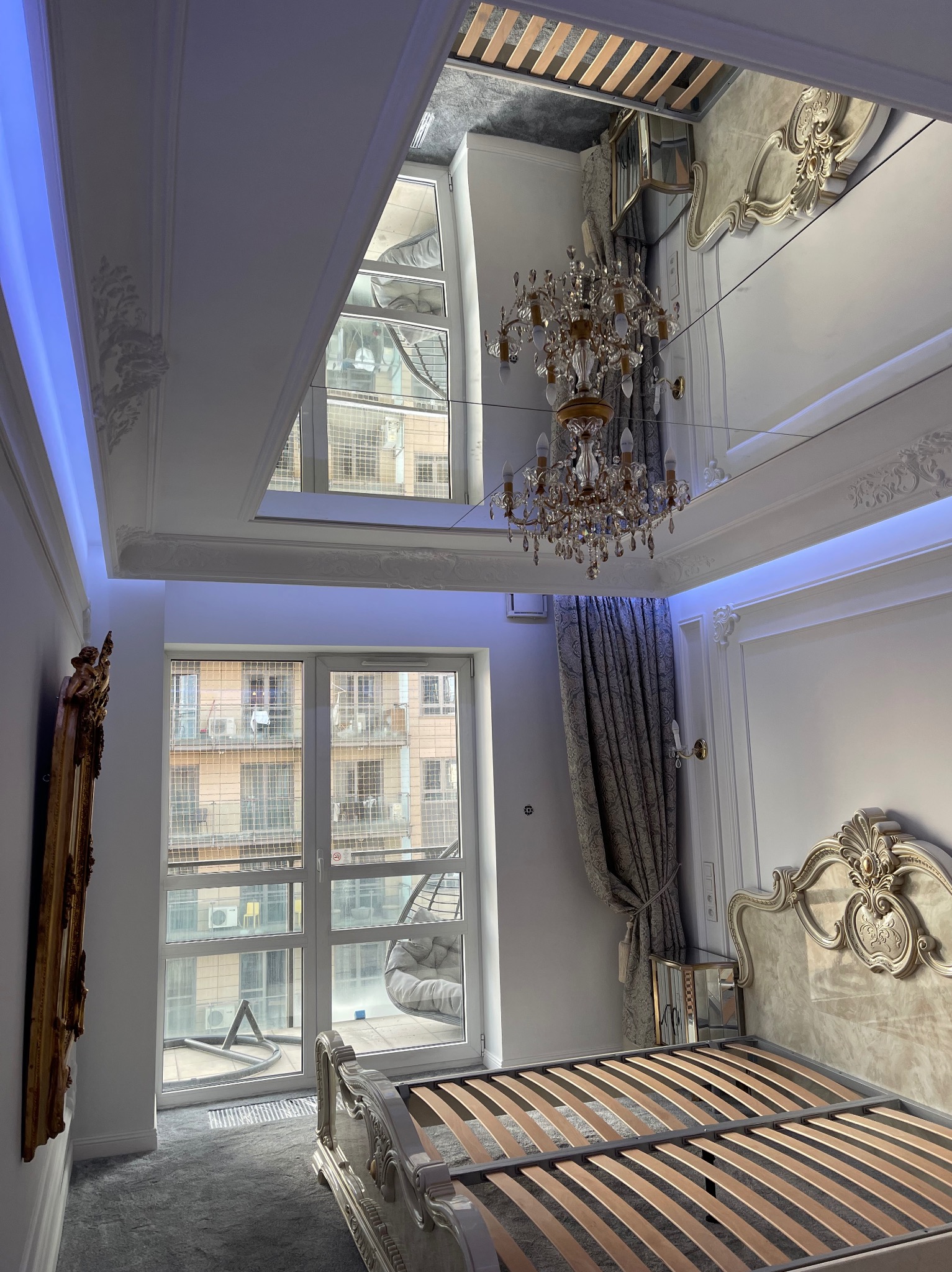 Elegancka sypialnia z lustrzanym sufitem, kryształowym żyrandolem i dekoracyjnym łóżkiem z rzeźbionym zagłówkiem, widok na balkon przez okno.