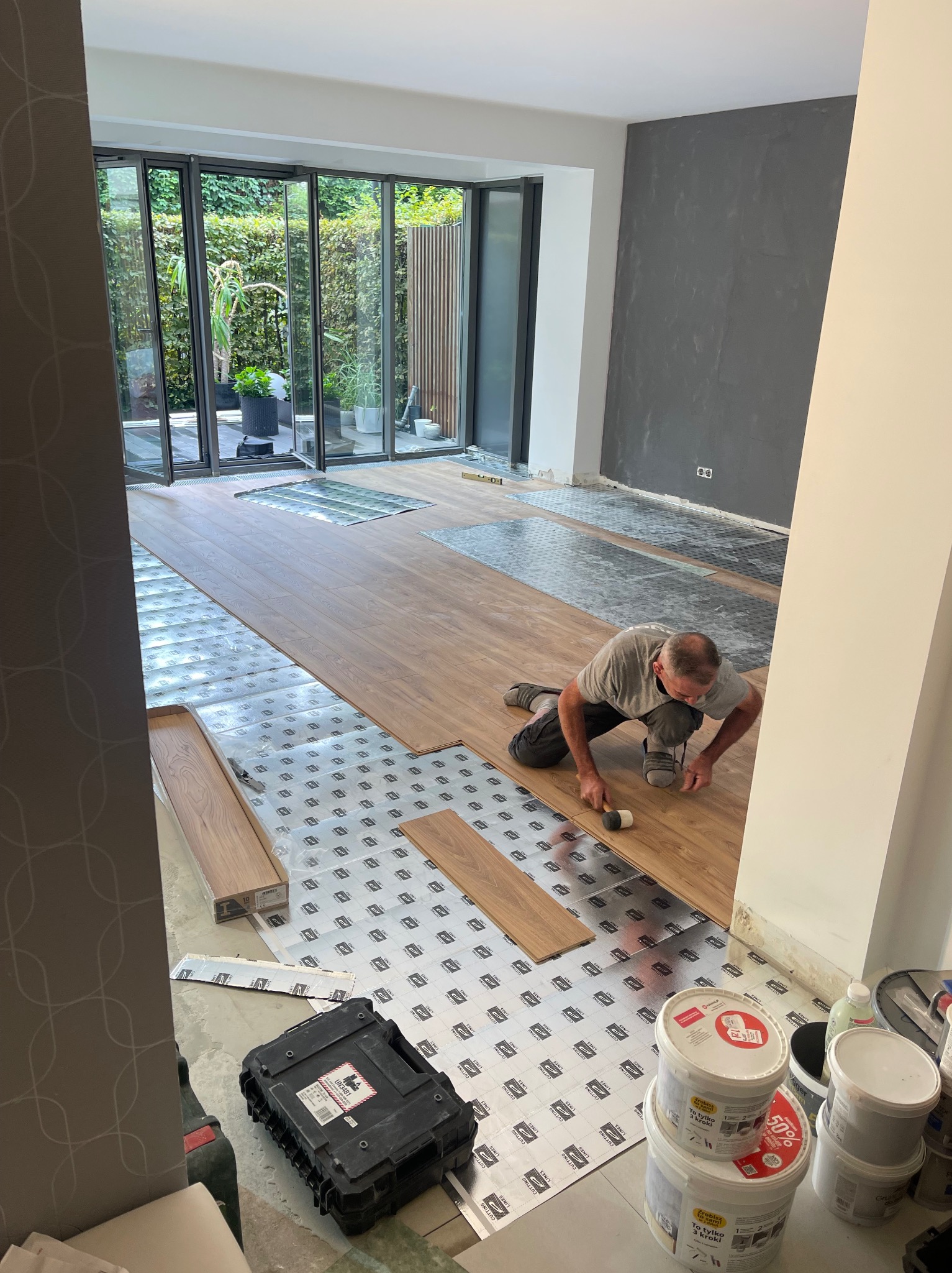 Układanie paneli podłogowych w pomieszczeniu z szarą ścianą akcentującą i dużymi oknami wychodzącymi na ogród; widoczny pracownik montujący panele na podkładzie izolacyjnym, narzędzia pracy...