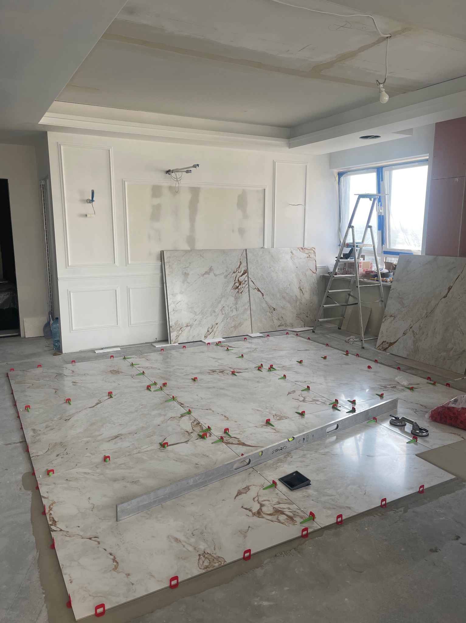 Układanie płytek imitujących marmur na podłodze w trakcie remontu, widoczne poziomica, krzyżyki dystansowe i zapasowe płytki oparte o ścianę.