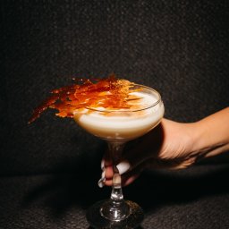 EXECUTIVE LOUNGE SPÓŁKA Z OGRANICZONĄ ODPOWIEDZIALNOŚCIĄ - Kremowy koktajl w kieliszku koktajlowym, ozdobiony kruchym karmelem i posypany przyprawami, trzymany w dłoni z białym manicure na tle ciemnej, teksturowanej ściany.