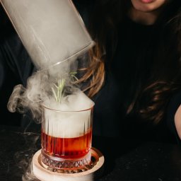 EXECUTIVE LOUNGE SPÓŁKA Z OGRANICZONĄ ODPOWIEDZIALNOŚCIĄ - Barman nalewa dym z szklanego naczynia do koktajlu Old Fashioned z gałązką rozmarynu, stojącego na drewnianej podstawce z ziarnami kawy.