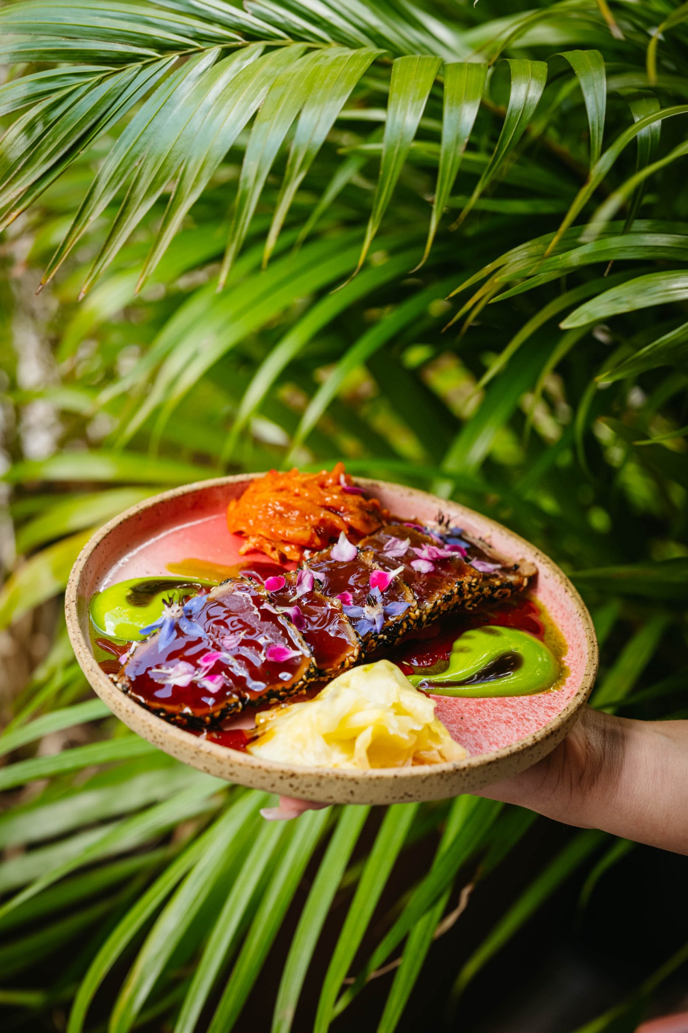 Elegancko podane danie z tuńczyka w sezamie, udekorowane jadalnymi kwiatami, z puree z batatów i imbirem, na tle tropikalnych liści palmowych.