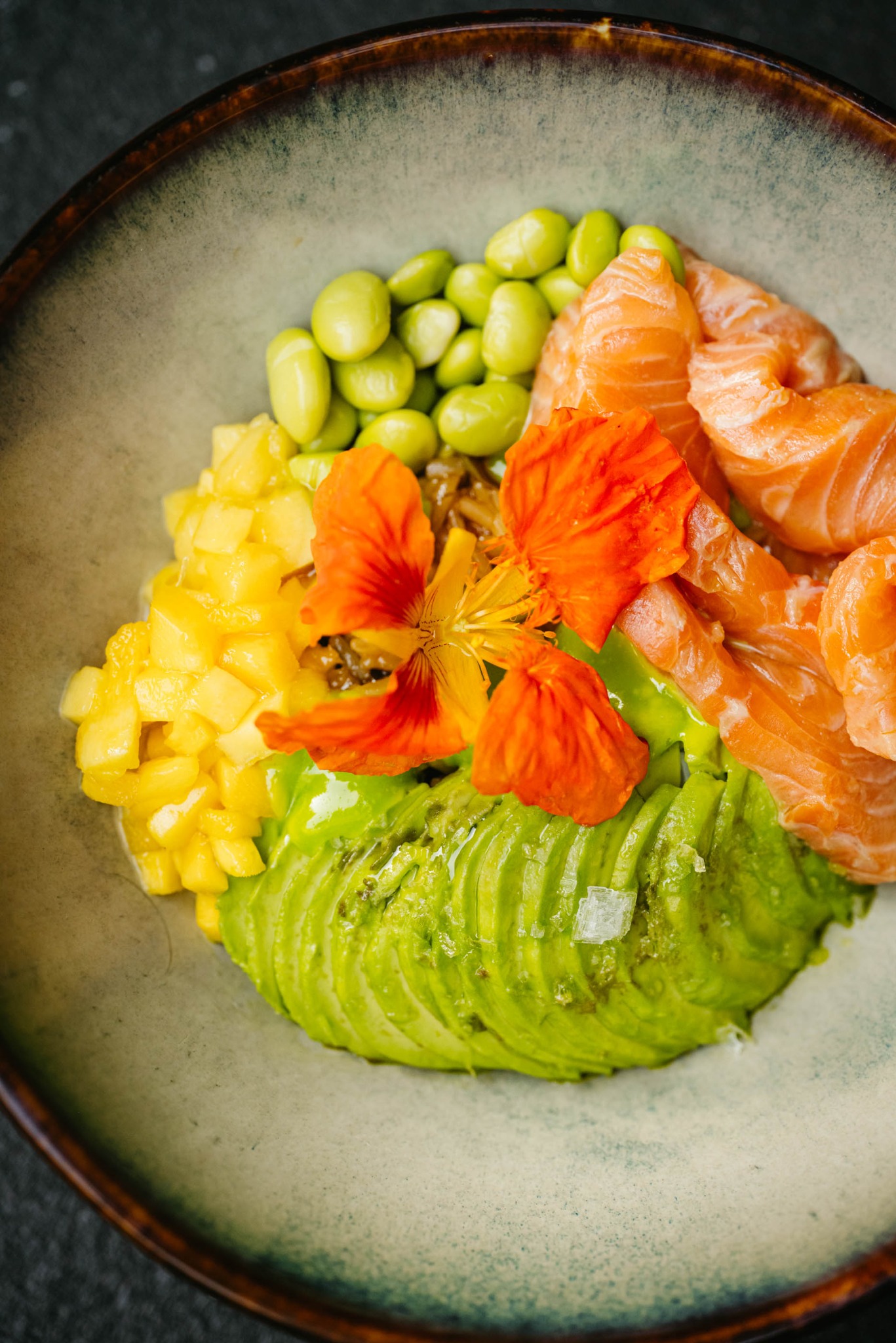 Artystycznie ułożona kompozycja poke bowl z łososiem, awokado, mango, edamame i jadalnym kwiatem nasturcji w ceramicznej misce z brązowym rantem, na ciemnym tle.