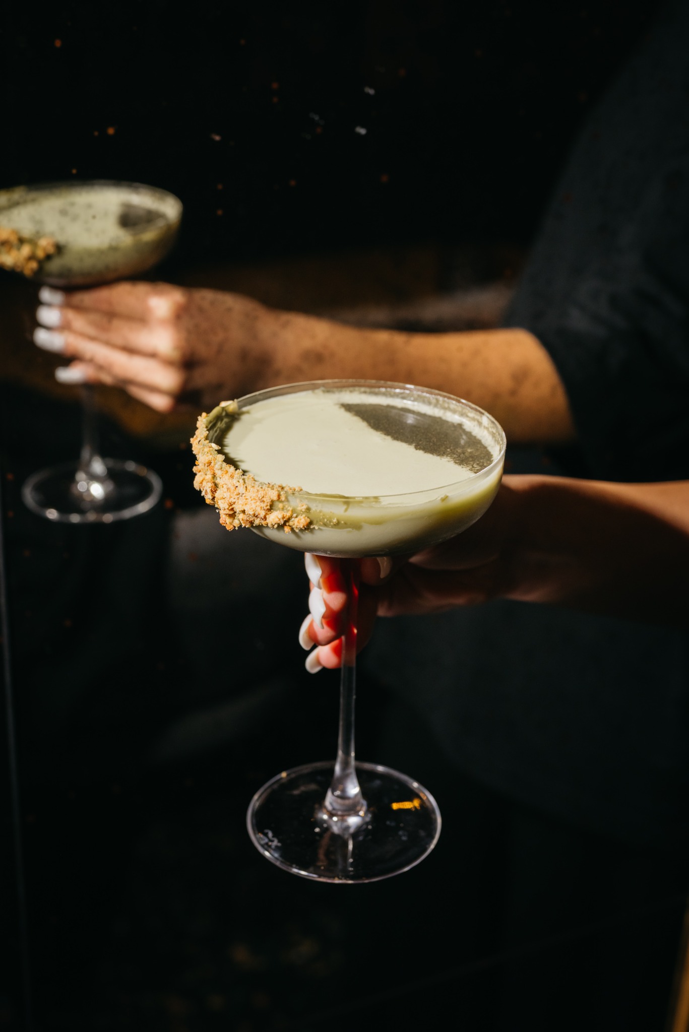 Dwa drinki w kieliszkach koktajlowych, z kremowym, zielonkawym napojem i posypką na krawędzi, trzymane przez kelnerki na ciemnym tle.