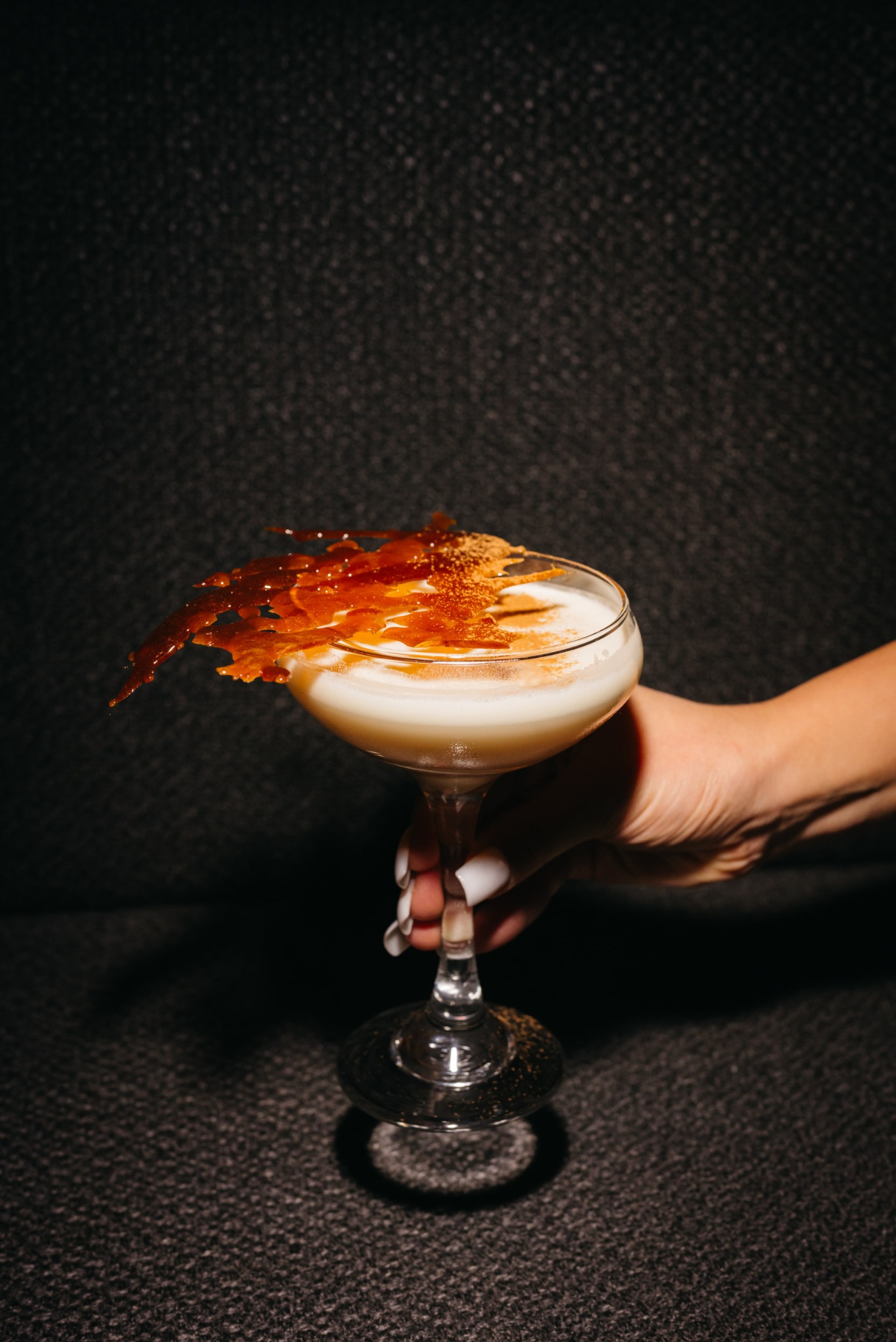 Kremowy koktajl w kieliszku koktajlowym, ozdobiony kruchym karmelem i posypany przyprawami, trzymany w dłoni z białym manicure na tle ciemnej, teksturowanej ściany.