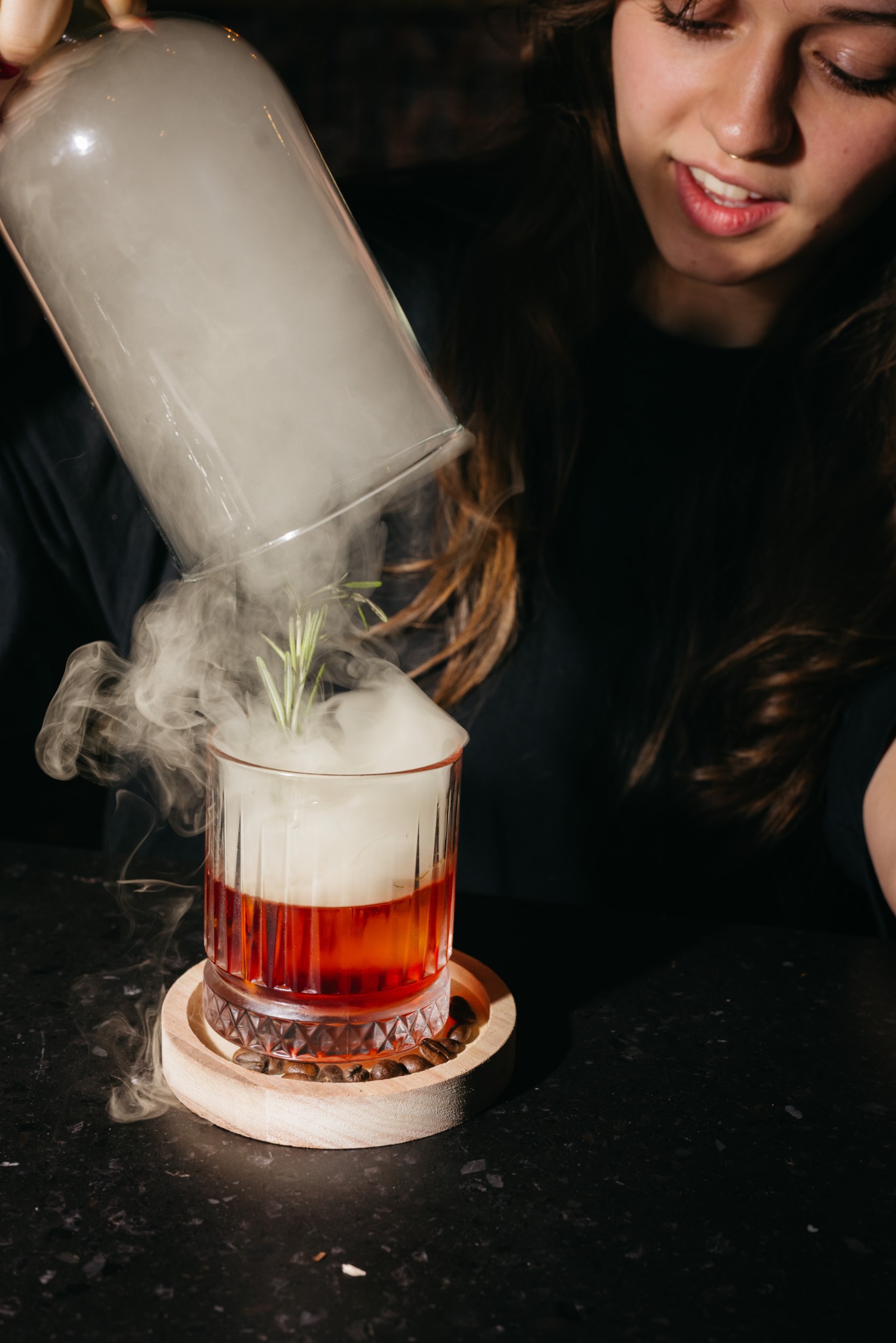 Barman nalewa dym z szklanego naczynia do koktajlu Old Fashioned z gałązką rozmarynu, stojącego na drewnianej podstawce z ziarnami kawy.