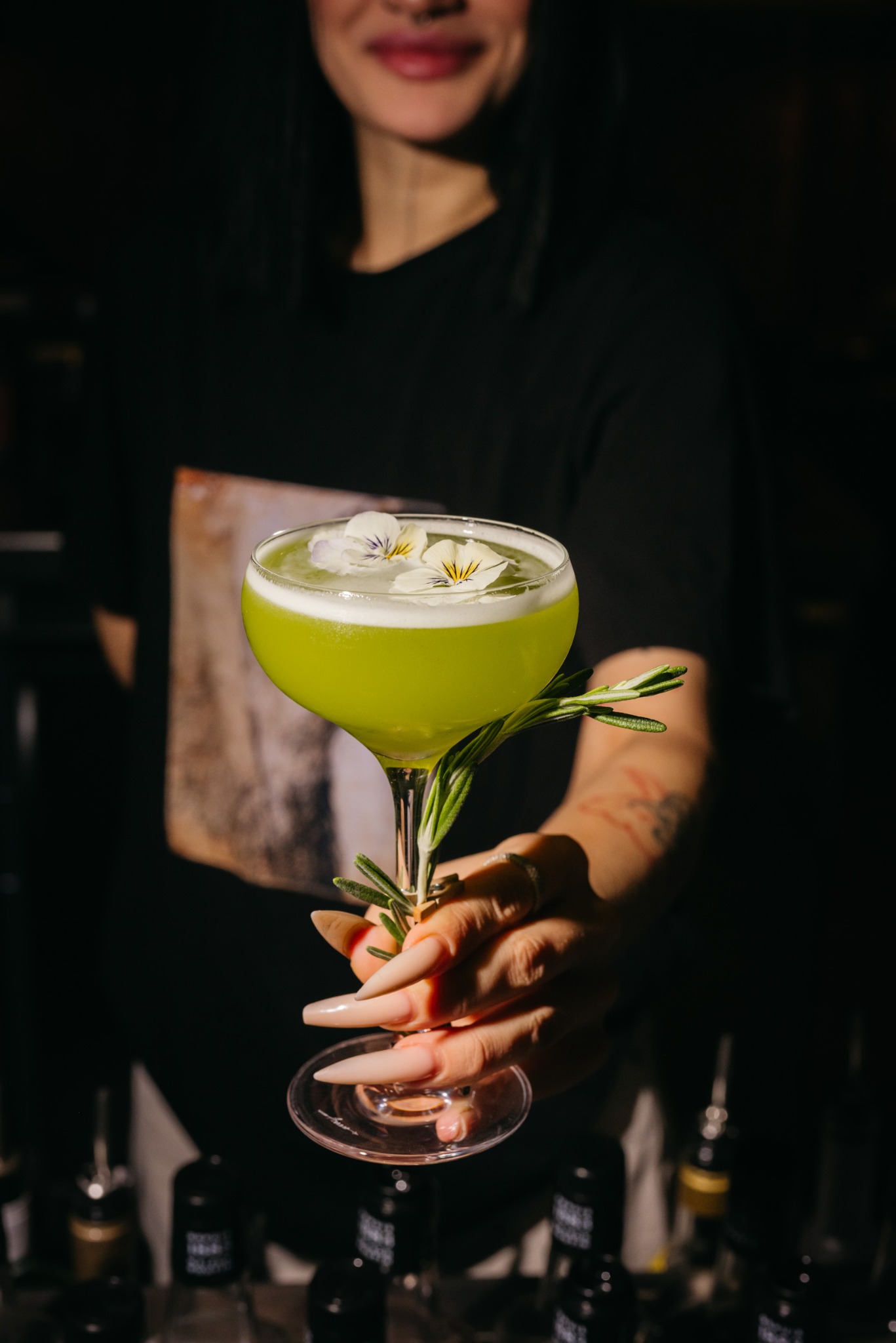 Kobieta trzyma w dłoni kieliszek z zielonym koktajlem, udekorowanym kwiatami i gałązką rozmarynu, na tle baru.