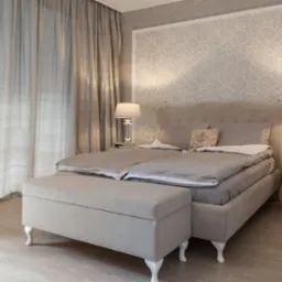 Elegancka sypialnia w odcieniach beżu i szarości z wzorzystą tapetą, lampkami nocnymi i łóżkiem z pojemnikiem, oświetlona naturalnym światłem przez delikatne zasłony.