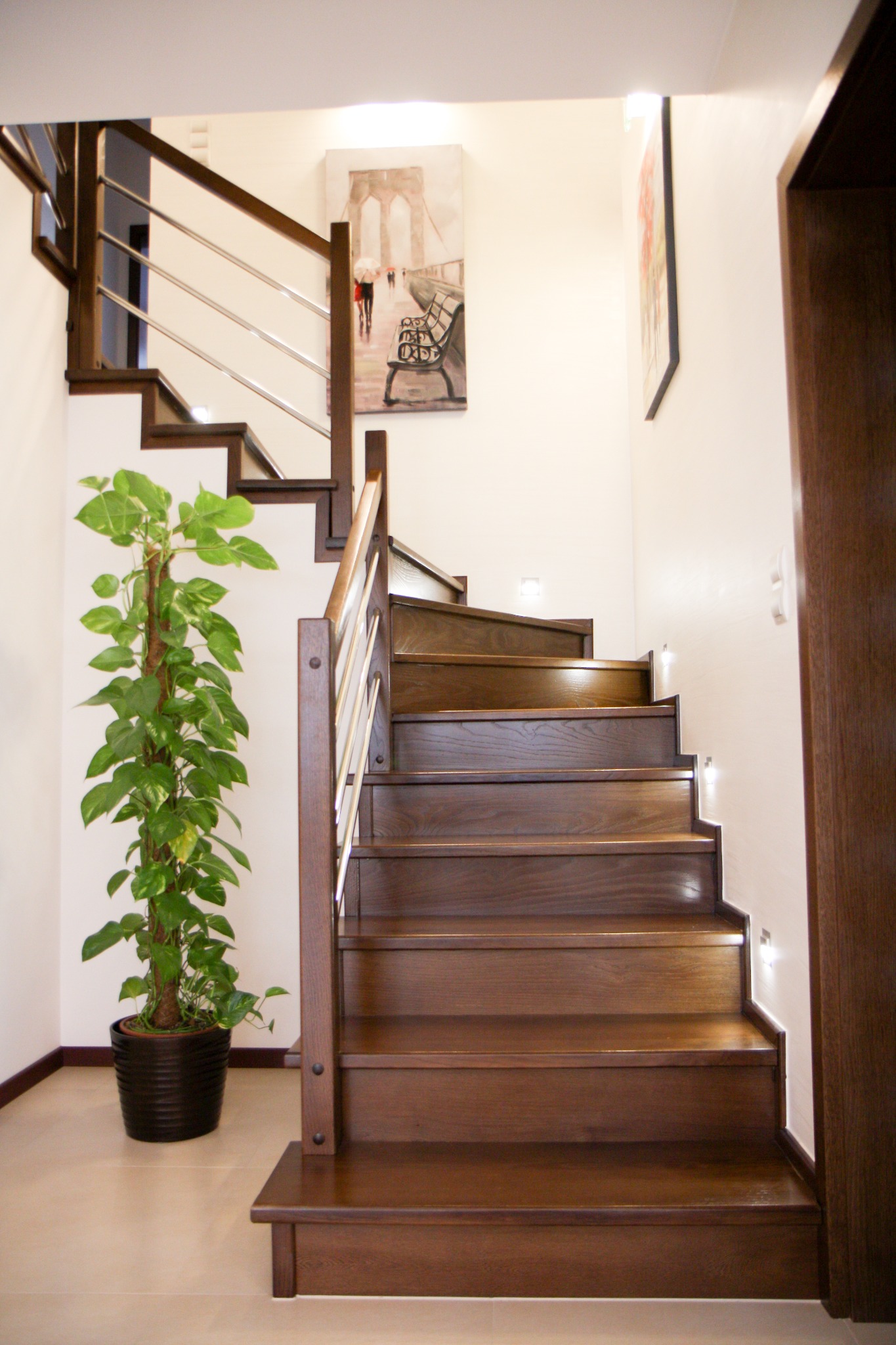 Schody z ciemnego drewna z metalową balustradą, oświetleniem punktowym na ścianie i rośliną doniczkową u podstawy, uzupełnione obrazem na ścianie.