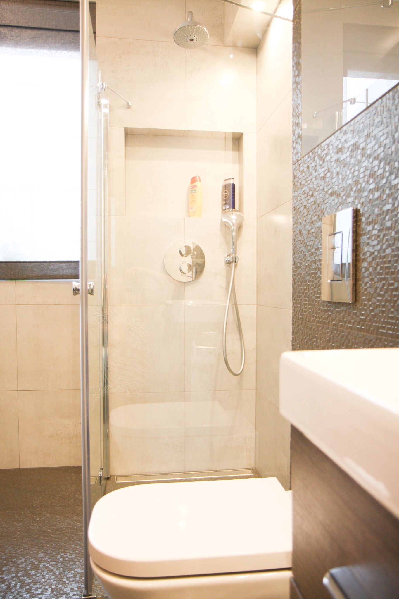 Nowoczesna łazienka z kabiną prysznicową typu walk-in, mozaikową ścianą, podwieszaną toaletą i wnęką z kosmetykami pod prysznicem.