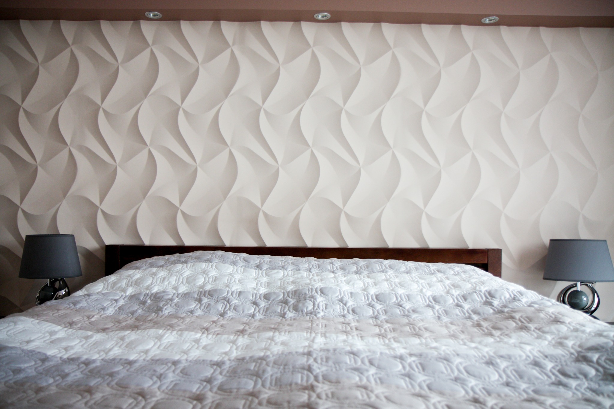 Sypialnia z dekoracyjną ścianą 3D w odcieniach beżu, brązowy zagłówek łóżka i biała pikowana narzuta, dwie szare lampki nocne po bokach.