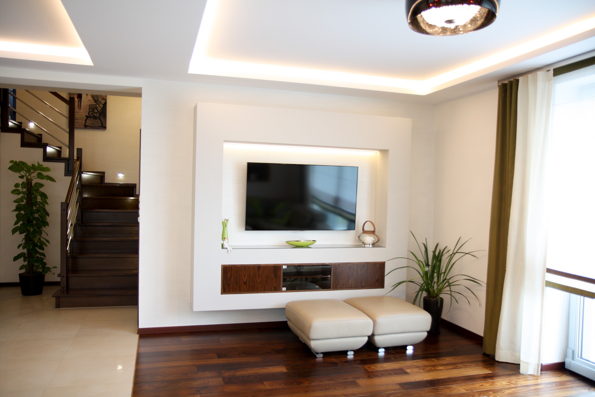 Nowoczesny salon z telewizorem w białej zabudowie, drewnianą podłogą i schodami w tle, oświetlenie LED w suficie i dekoracyjne dodatki.