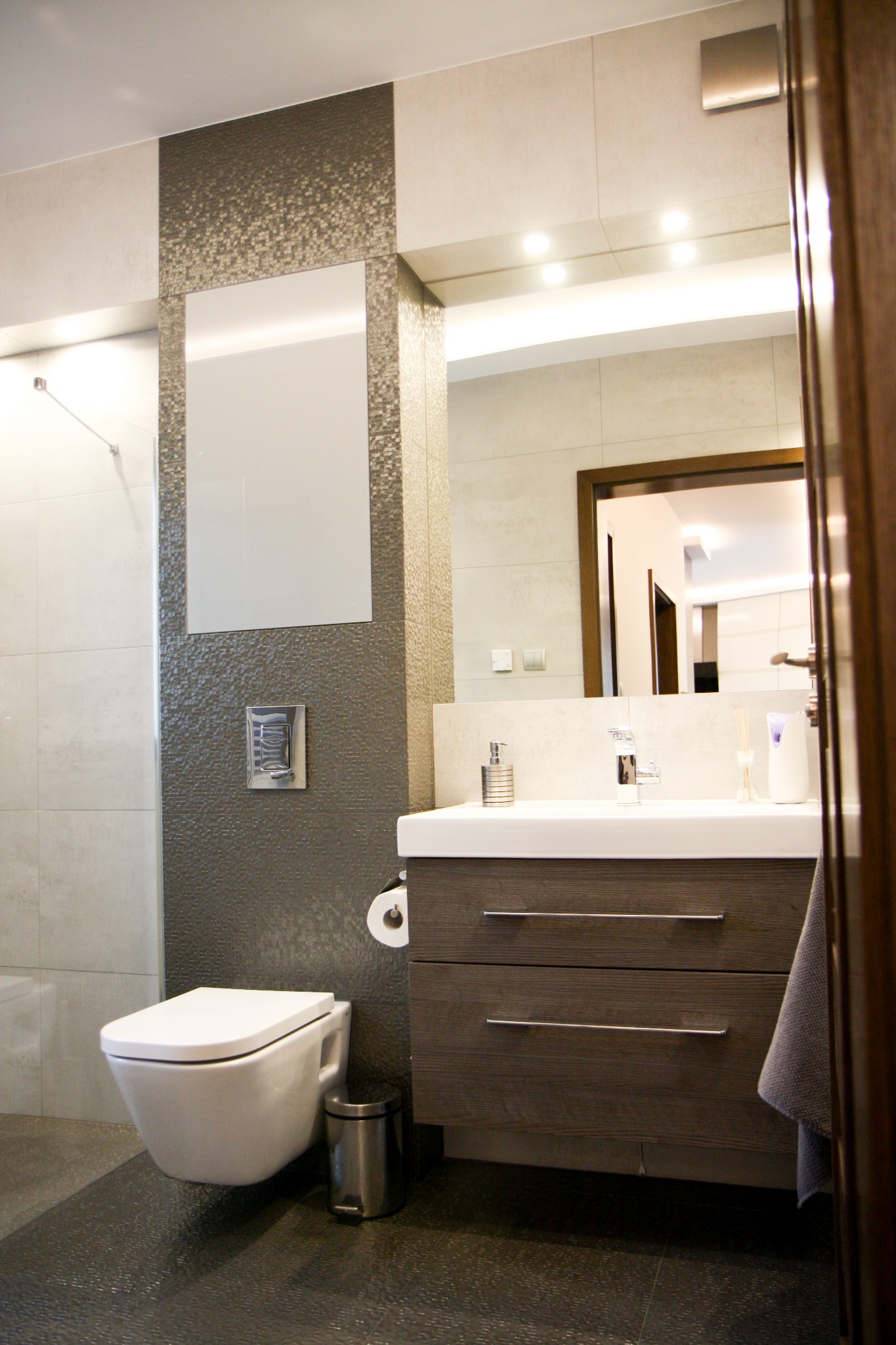 Nowoczesna łazienka z podwieszaną toaletą, umywalką z szafką w odcieniach szarości i beżu, oraz lustrem z oświetleniem LED, widok częściowo zasłonięty przez otwarte drzwi.