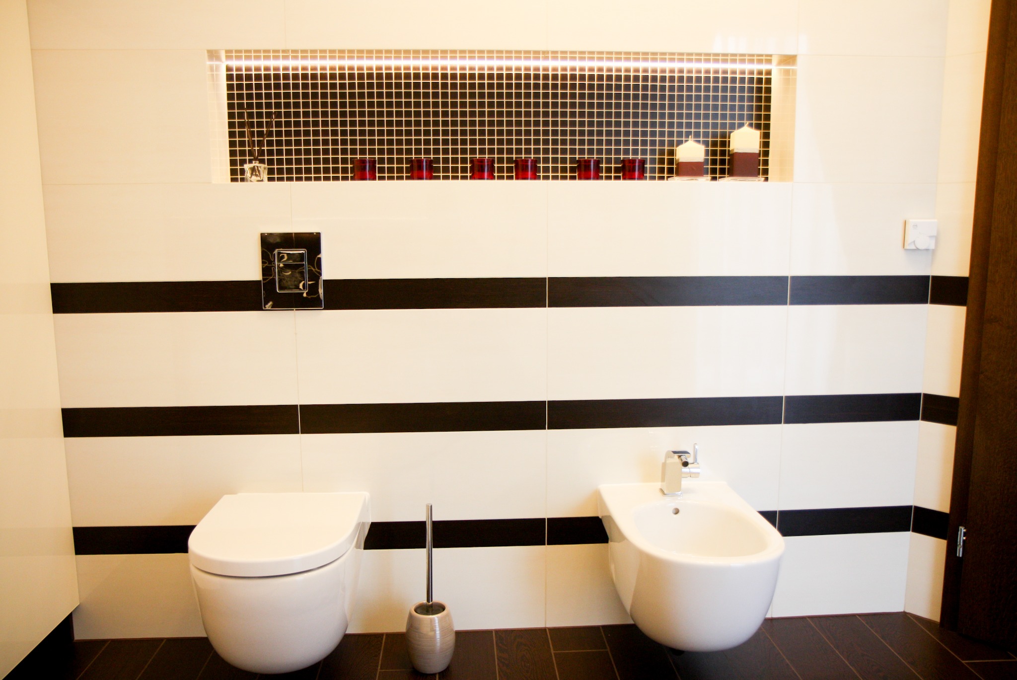 Nowoczesna łazienka z białymi płytkami w czarne pasy, wiszącą toaletą, bidetem i dekoracyjną wnęką z mozaiką, podświetleniem LED i ozdobnymi świeczkami.