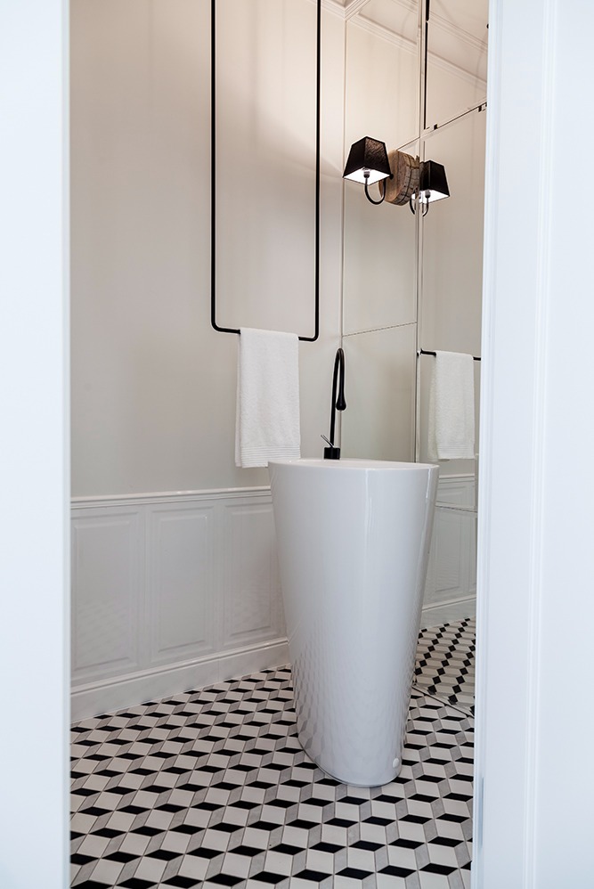 Nowoczesna łazienka z białą, wolnostojącą umywalką na tle ściany z panelami i podłogi w geometryczny wzór, oświetlona lampami z abażurami.