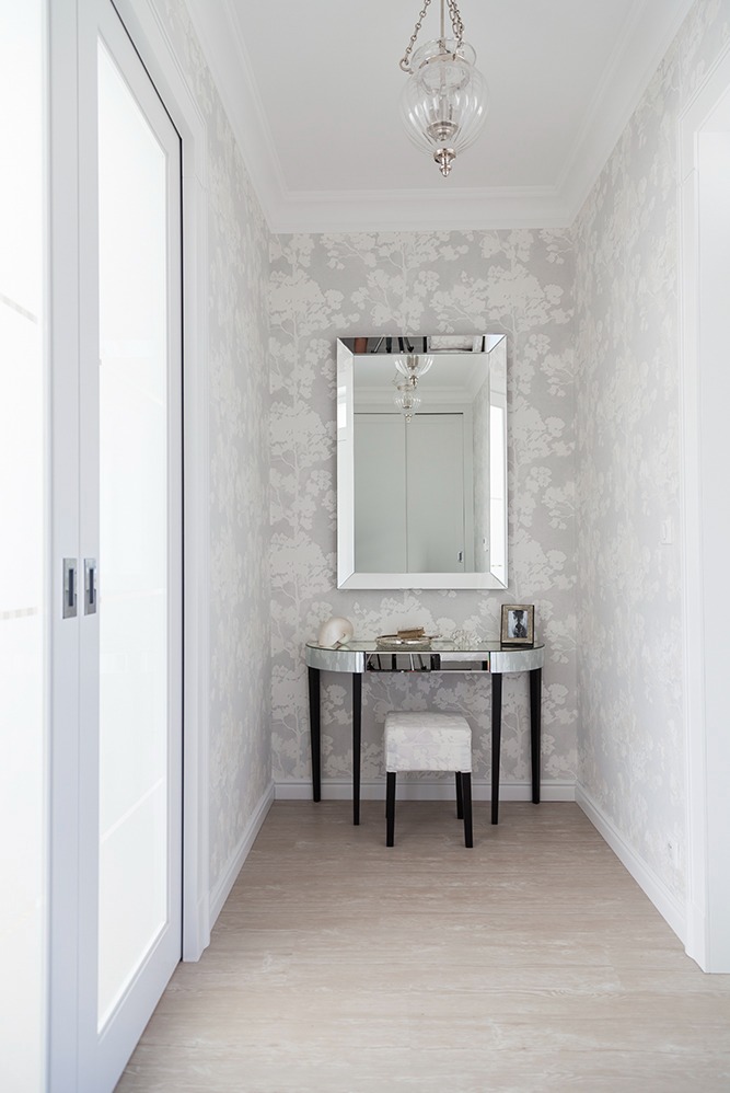 Wnętrze przedpokoju z tapetą w delikatny wzór drzew, lustrzana toaletka z krzesłem i designerska lampa wisząca.