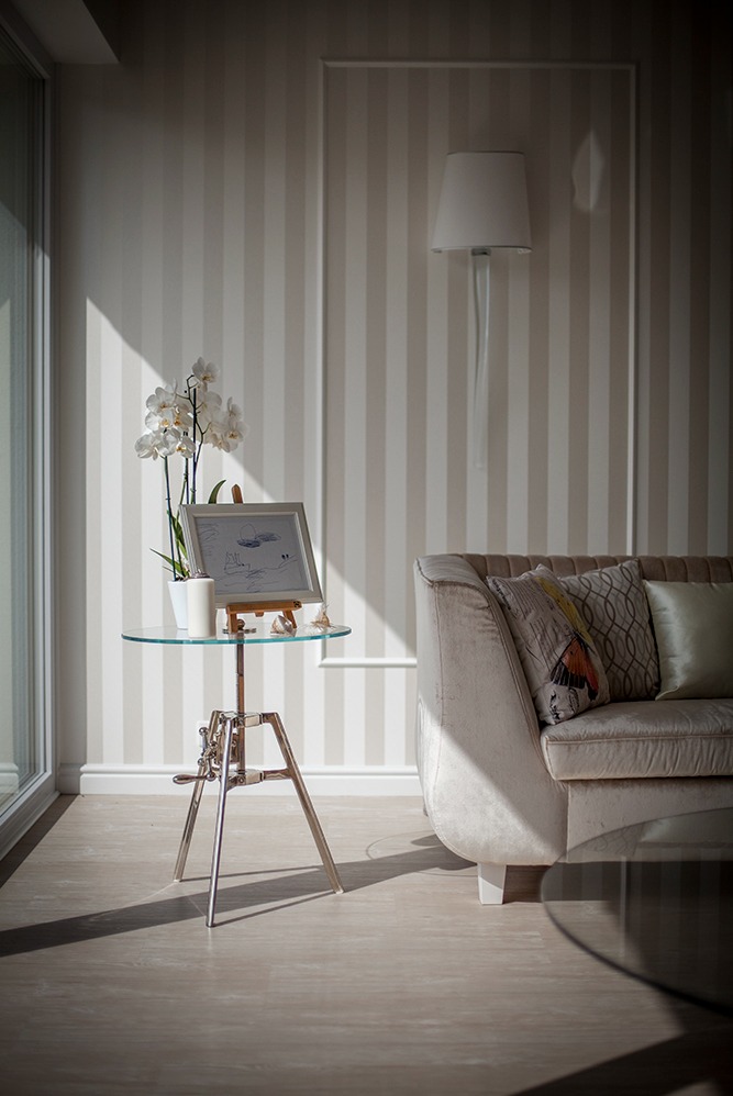 Eleganckie wnętrze pokoju z tapetą w pionowe pasy, szklanym stolikiem na metalowym statywie, kwiatami orchidei w doniczce i beżową sofą z dekoracyjnymi poduszkami, oświetlone promieniami słońca.