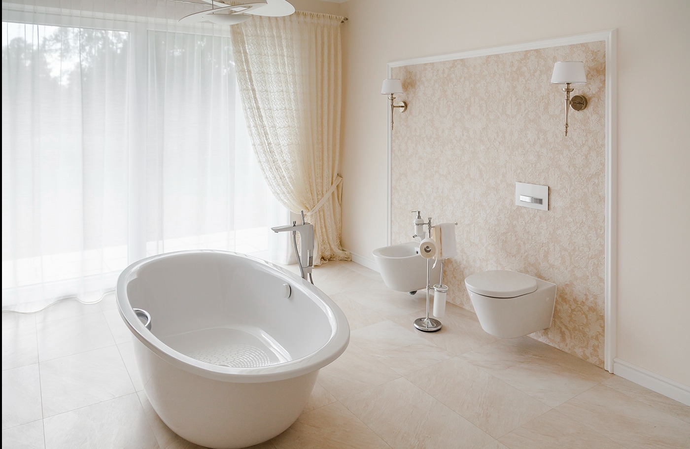Jasna łazienka z wolnostojącą wanną, bidetem i wiszącą toaletą na tle ściany w delikatny wzór, oświetlona kinkietami i naturalnym światłem z okna z firanką.
