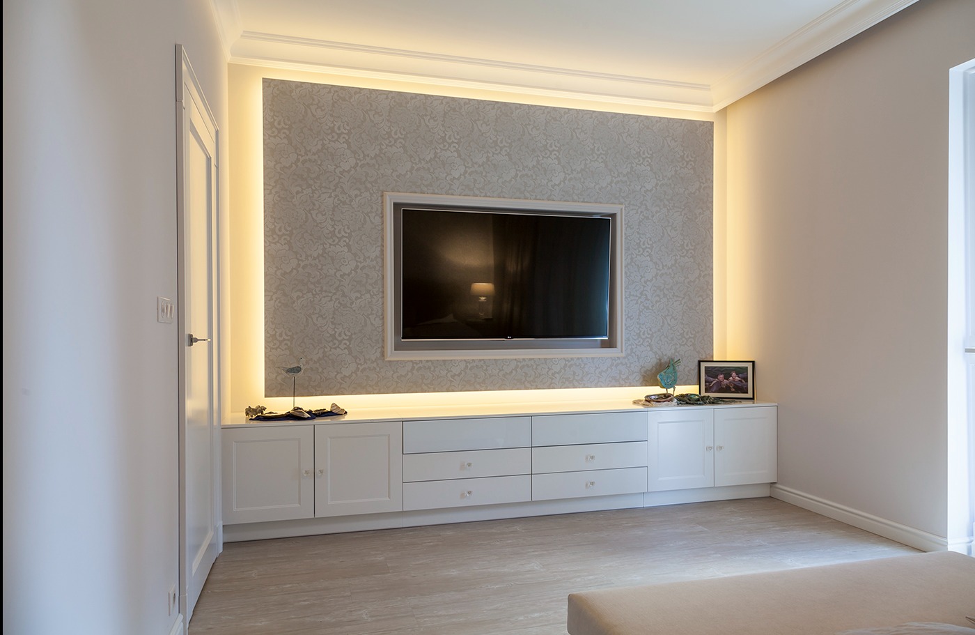 Elegancki salon z zabudową meblową pod telewizorem, podświetlonymi wnękami i wzorzystą tapetą w odcieniach beżu.
