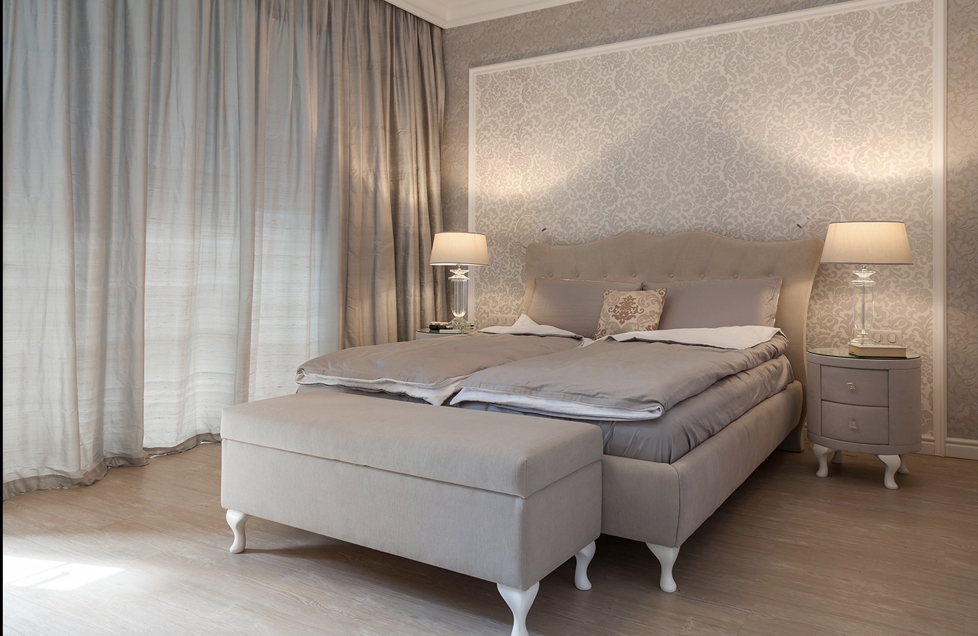 Elegancka sypialnia w odcieniach beżu i szarości z wzorzystą tapetą, lampkami nocnymi i łóżkiem z pojemnikiem, oświetlona naturalnym światłem przez delikatne zasłony.